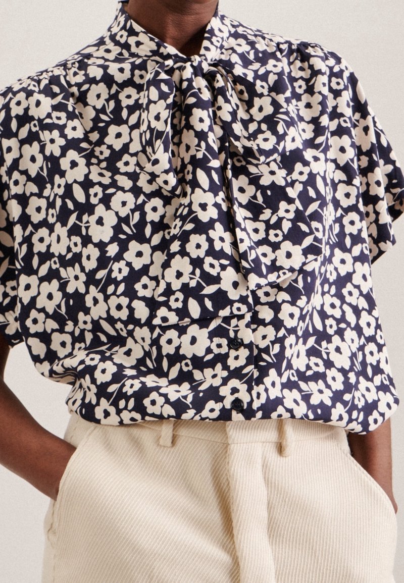 Blommig blus i marinblått tyg med vita blommor, med en rosettkrage och kortärmar. Kombinerad med ljusa, texturerade byxor.