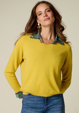 Donna con lunghi capelli castani indossa grandi orecchini, un maglione giallo sopra una camicia blu a fantasia e jeans blu, in piedi con le mani nelle tasche.