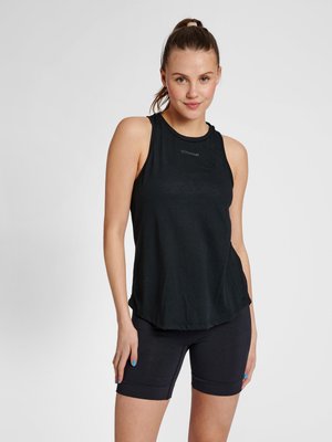 Jonge vrouw met paardenstaart die een zwarte mouwloze sporttop en zwarte aansluitende shorts draagt, staand tegen een egale lichte achtergrond.