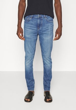 Man draagt slim-fit blauwe spijkerbroek, zwart T-shirt en zwarte open-toe instapschoenen, staand tegen een effen witte achtergrond.