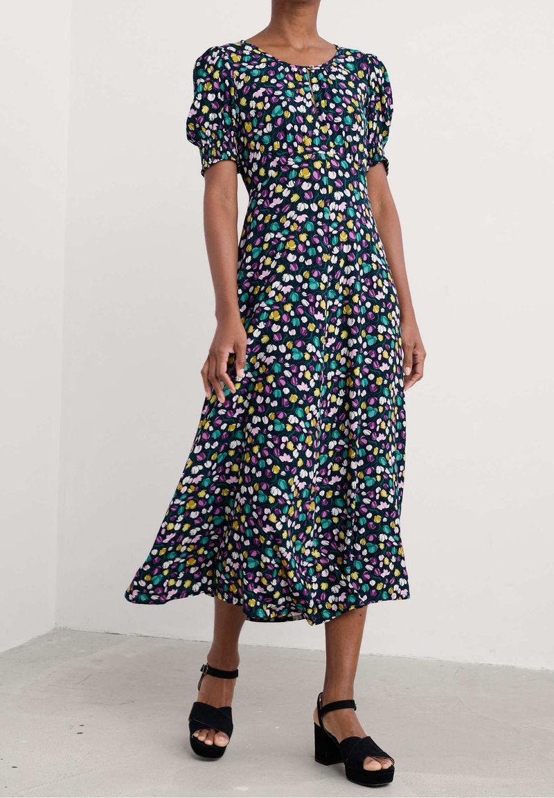 Femme portant une robe midi bleu marine à manches bouffantes et un motif floral abstrait multicolore, associée à des sandales compensées noires.