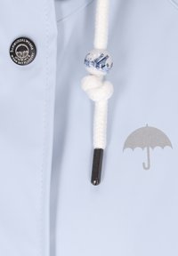 Šviesiai mėlyna vandeniui atspari striukė su metaliniu logotipo mygtuku, išsiuvinėtu sidabro spalvos skėčio detaliu ir dekoratyviniu mazgu su ornamentuotu karoliuku.