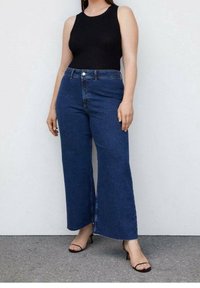 Jeans de mezclilla azules de tiro alto y pierna ancha con una textura suave, bolsillos frontales y cierre de botón. Combinados con una blusa negra sin mangas ajustada.