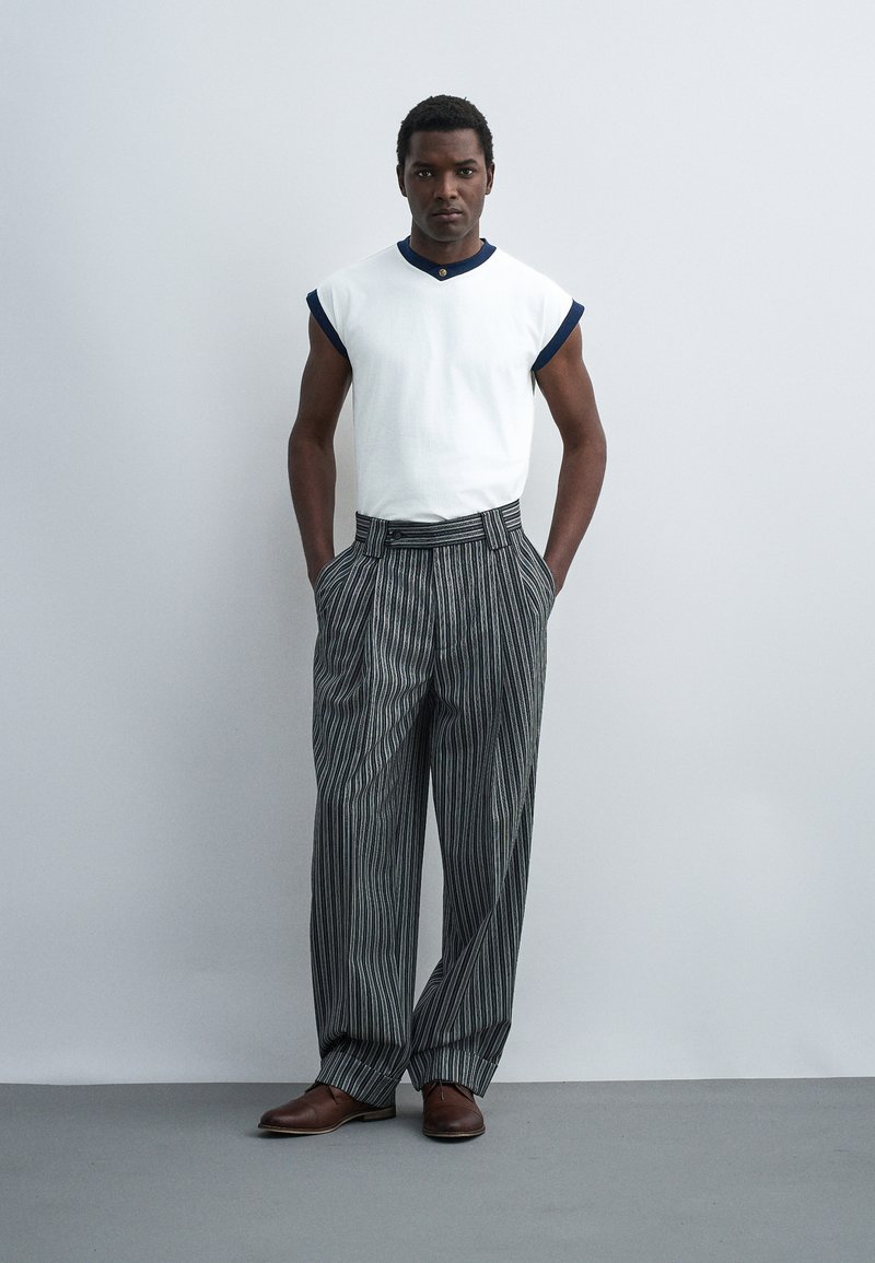 Young man standing with hands in pockets, wearing a white sleeveless shirt with navy trim, wide gray pinstriped trousers, and brown shoes.