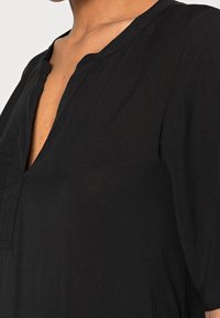 Gros plan d'une personne portant une chemise noire à col en V avec des manches trois-quarts, montrant une partie du cou et du haut de la poitrine.