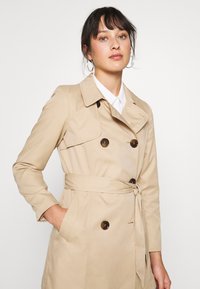 Beige trenčkot s dvojitým zapínáním, velkými hnědými knoflíky, opaskem v pase a klopovým límcem; hladká textura látky.