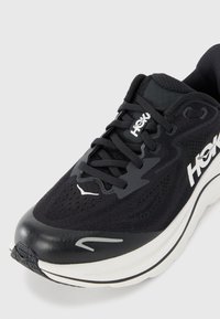 Schwarzer Sportschuh mit einem atmungsaktiven Mesh-Obermaterial, gepolstertem Schaft, dicker weißer Zwischensohle und schwarzem Außensohle, mit HOKA-Branding.