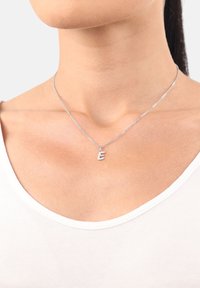 Collana d'argento con un ciondolo a forma di "e" minuscola, caratterizzata da una catena delicata e una superficie liscia, indossata sopra una semplice camicia bianca.