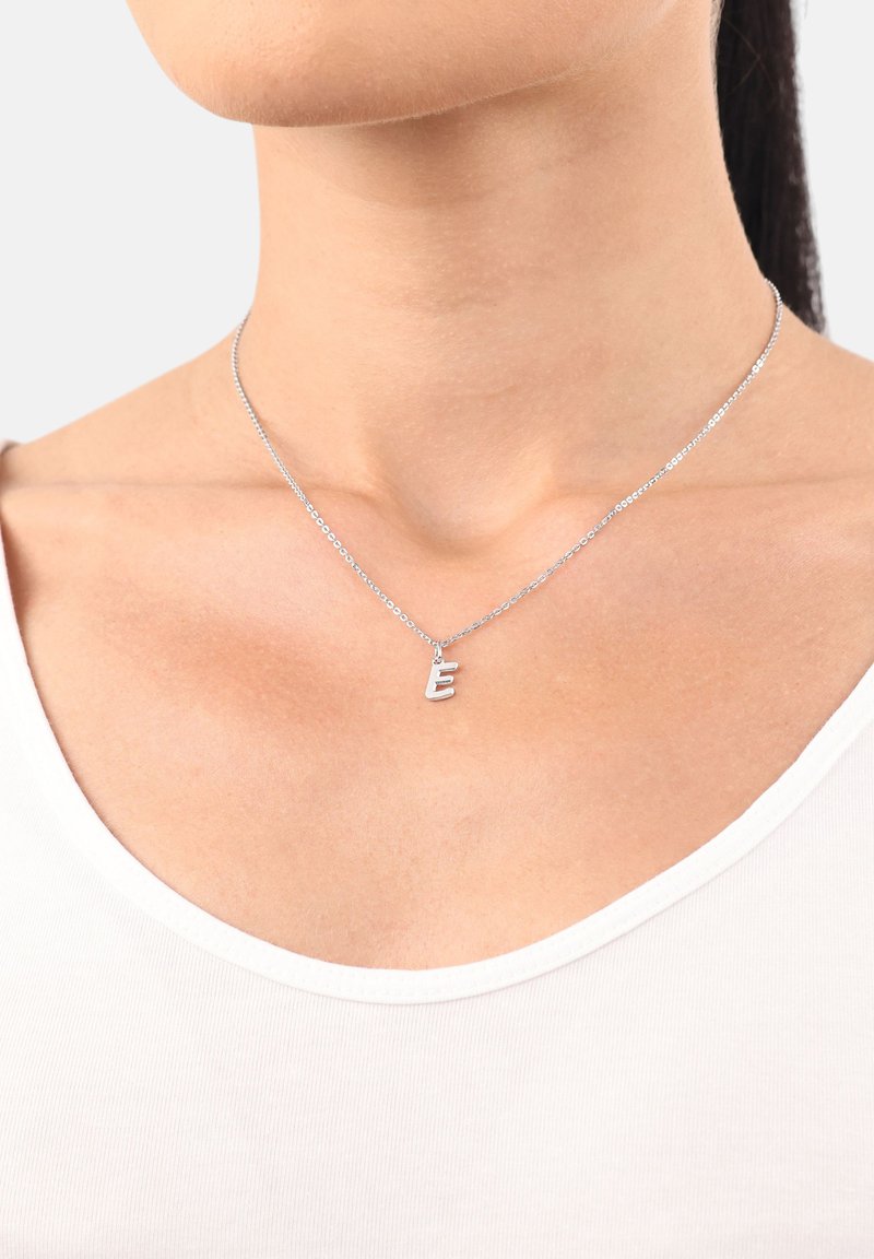 Collana d'argento con un ciondolo a forma di "e" minuscola, caratterizzata da una catena delicata e una superficie liscia, indossata sopra una semplice camicia bianca.