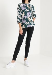 Blommig blus i mörkgrön med vita och lila blommor, rund halsringning, 3/4 långa ärmar; matchad med svarta skinny jeans och vita sneakers.