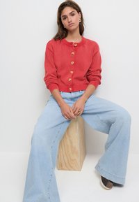 Red knitted cardigan with wooden buttons, ribbed cuffs, and a round neckline. Paired with light blue wide-leg jeans and brown shoes.
