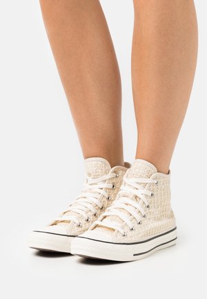 Wysokie trampki z jasnobeżowej tkaniny plecionej, z białymi sznurowadłami, metalowymi przelotkami i gumową podeszwą z czarnymi paskami.