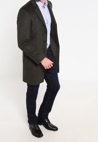 Long manteau vert foncé avec un col cranté, fermeture à boutons et poches latérales, porté sur une chemise à carreaux bleu clair et un jean sombre avec des chaussures noires.