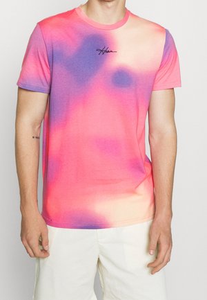 Mężczyzna ubrany w krótkim rękawem koszulkę tie-dye w kolorach różowym, fioletowym i beżowym, w połączeniu z białymi spodniami, na jednolitym tle.