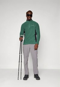 Zöld polár cipzáras felső fekete oldalsó betétekkel, szürke nadrág és fekete túracipő. Túrabotokkal a kezében; napszemüveg teszi teljessé a szettet.