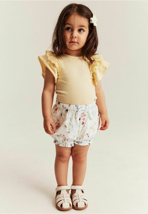 Ung pige iført en gul top med flæser på ærmerne, hvide blomstrede shorts, hvide sandaler og en hvid hårclips, stående foran en ensfarvet baggrund.