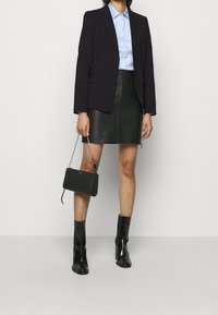 Kvinna klädd i en svart blazer, blå skjorta med knappar, svart läder minikjol, svarta ankelskor, hållande en liten svart handväska med kedjesnodd.