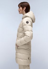 Abrigo acolchado beige con capucha, que presenta un diseño acolchado, bolsillos con cremallera y un parche con logo en el hombro. Textura suave y corte de longitud media.