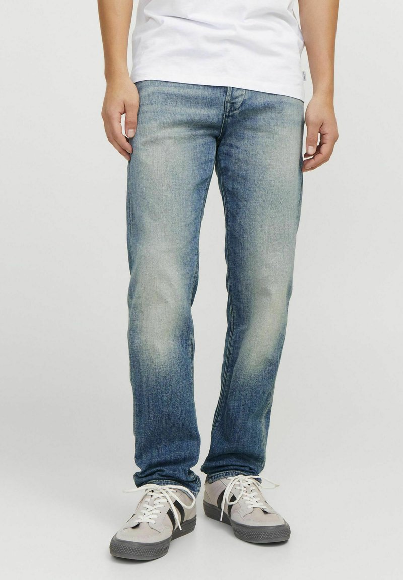 Vaqueros de mezclilla azul deslavado con corte recto, presentan un sutil desgaste, cinco bolsillos y un lavado medio. Combinados con zapatillas grises.