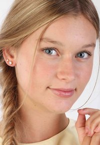 Studs met heldere edelstenen, gezet in roségoud. Glad oppervlak en ronde vormen met drie stenen op elke oorbel, gedragen door een lichtgekleurd persoon.