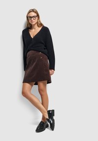 Zwarte fuzzy cardigan met een v-hals. Bruine corduroy wikkelrok met twee ronde knopen. Zwarte platform loafers. Neutrale achtergrond.