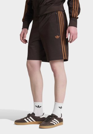 Męski model noszący ciemnobrązowe spodenki Adidas z pomarańczowymi paskami i logo, zestawione z białymi skarpetkami Adidas i ciemnobrązowymi butami sportowymi.