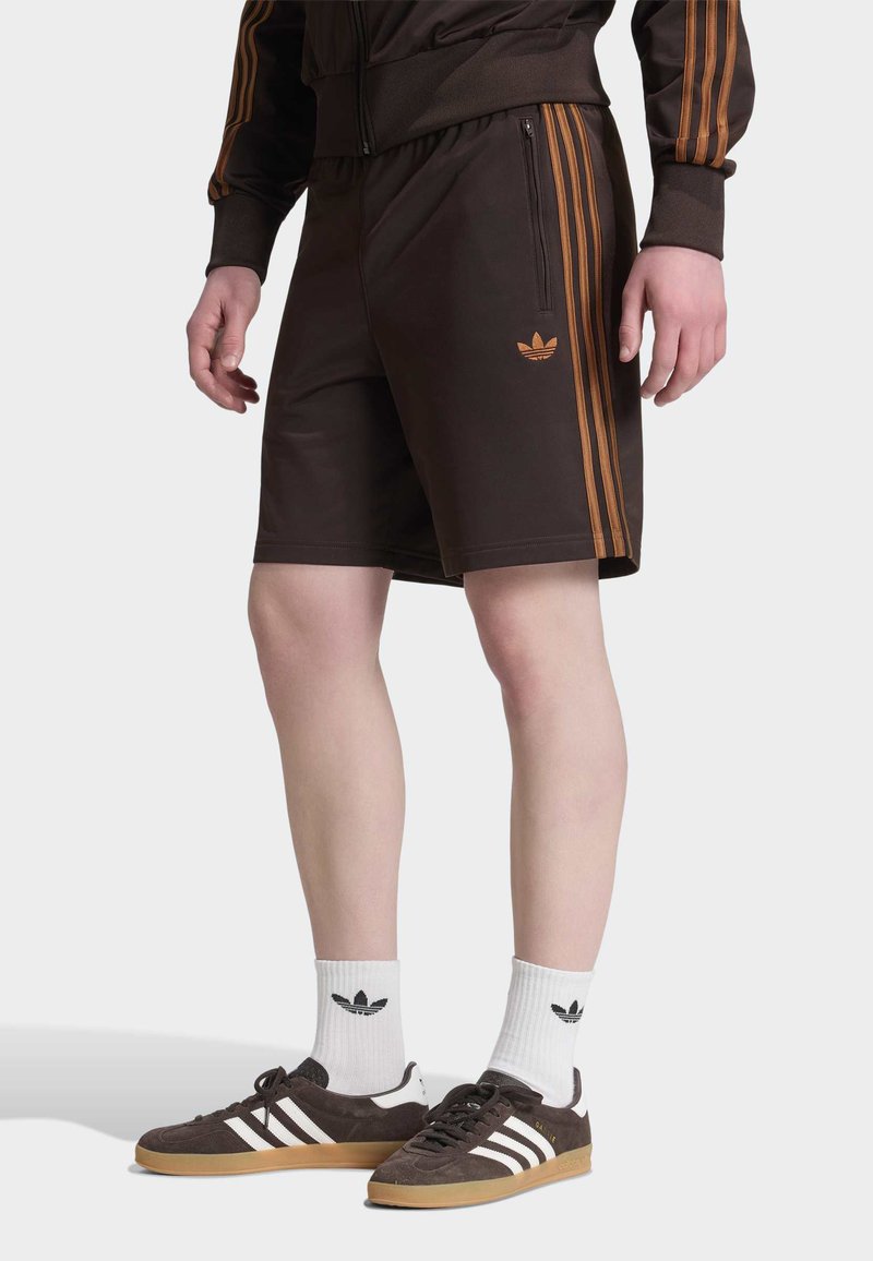 Άνδρας μοντέλο φοράει σκούρα καφέ σορτς Adidas με πορτοκαλί ρίγες και λογότυπο, συνδυασμένα με λευκές κάλτσες Adidas και σκούρα καφέ αθλητικά παπούτσια.