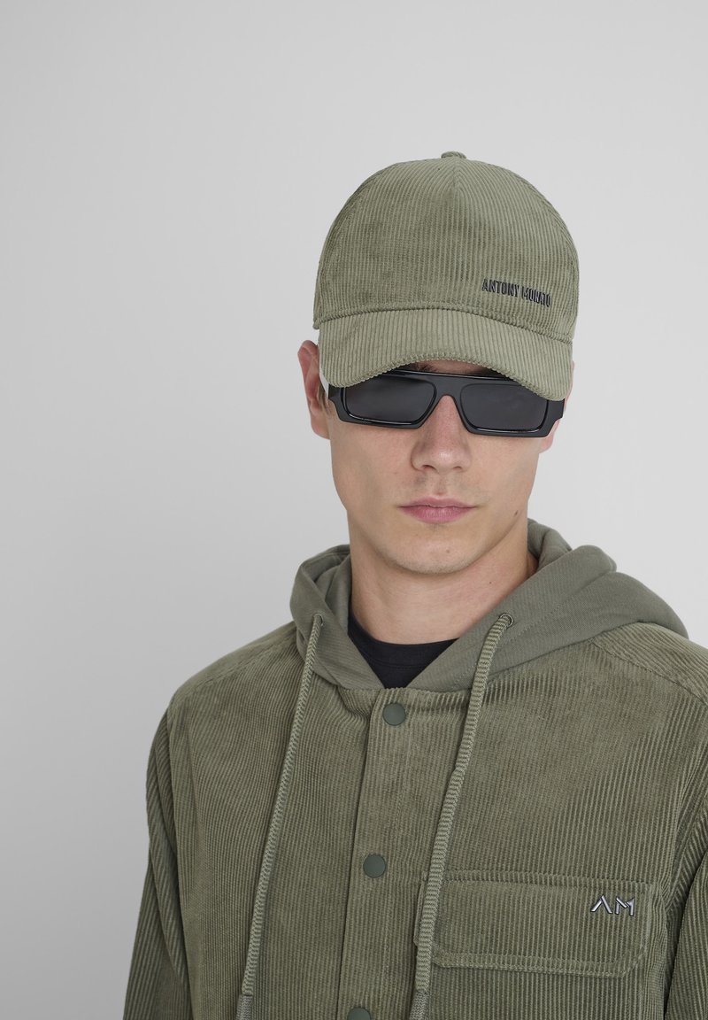 Gorra de canalé verde oliva con un acabado texturizado, combinada con una chaqueta de canalé a juego que presenta una capucha y detalles de botones.