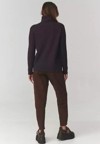 Kobieta z długimi włosami nosi ciemny prążkowany sweter z golfem, brązowe spodnie i czarne buty na platformie, stojąc na jednolitym jasnym tle.