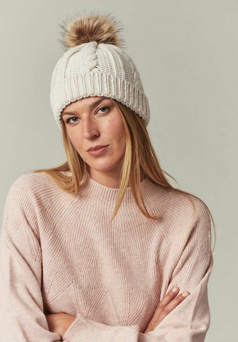 Crème gebreide beanie met een bont pompon bovenop; gemêleerd, geribbeld patroon; gecombineerd met een lichtroze, geribbelde trui met een chevron ontwerp.