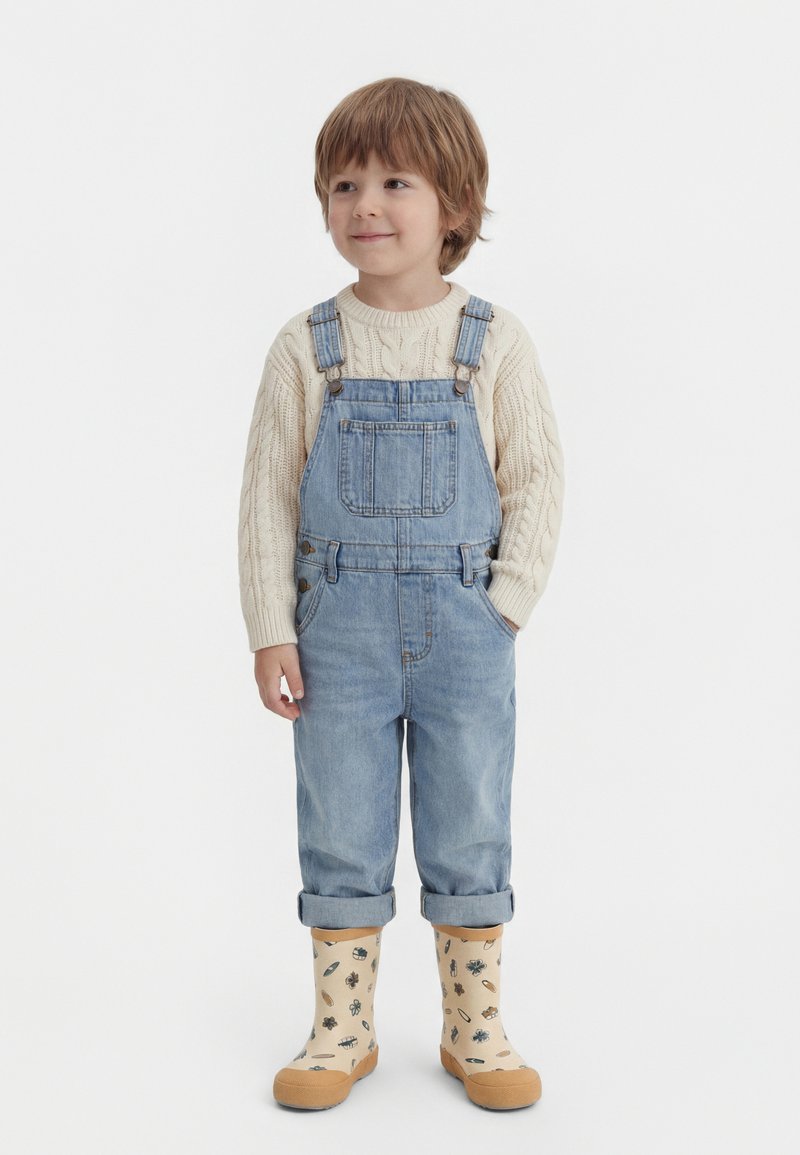 Fiatal gyermek világosbarna hajjal, bézs kötött pulóvert, kék farmer kantáros nadrágot és krémszínű esőcsizmát visel, amelyeken természetminták vannak, és zsebében tartott kezekkel áll.