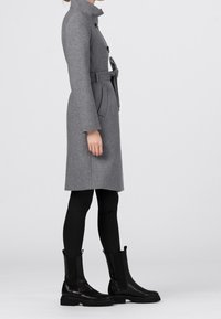 Grå ullrock med hög krage, knappstängning fram, bälte i midjan och sidofickor, kombinerad med svarta läderstövlar vid anklarna och snäva svarta byxor.