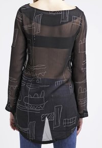 Blouse noire transparente à manches longues avec des motifs abstraits blancs. Ourlet arrière légèrement allongé, décolleté rond et texture douce.
