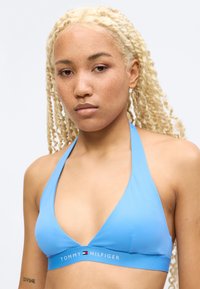 Νεαρός με ξανθά, πλεγμένα μαλλιά και φακίδες φοράει μπλε τοπ μπρά που δένει στον λαιμό Tommy Hilfiger, σε λευκό φόντο.