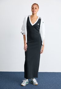 Robe maxi noire à côtes avec un décolleté en V, veste de sport blanche et baskets argentées avec des accents verts. Présentée sur un fond uni.