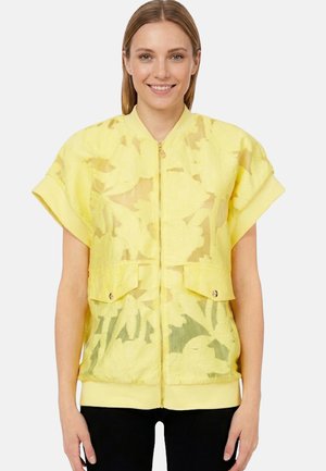 Donna sorridente con capelli lisci che indossa una giacca gialla con zip e motivo floreale a maniche corte e pantaloni neri su sfondo bianco.