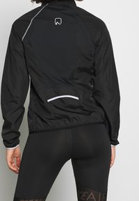Zwarte windjack met een gaaspaneel aan de achterkant, reflecterende accenten en een aansluitend ontwerp. Bevat een klein logo op de bovenrug.