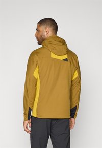 Hættejakke i letvægtsmateriale i jordbrun og gul med en struktureret overflade. Har kontrastdetaljer og et sporty design.