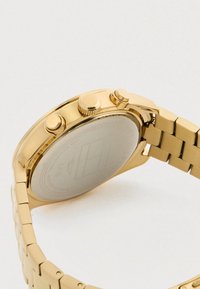 Kuldne käekell metallist lingirihmaga, millel on näha tagakate ja külgne nupud, jäädvustatud lihtsal valgel taustal.