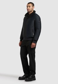 Musta bomber-takki, jossa on teksturoitu, turkista vuorattu kaulus, vetoketjulliset taskut, uritettujen hihansuiden kanssa ja sileä pinta, yhdistettynä mustiin cargo-housuihin ja saappaisiin.