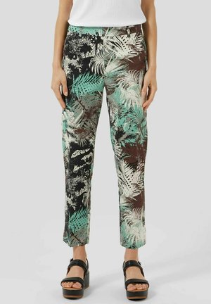 Kobieta w dopasowanych spodniach w tropikalny wzór w odcieniach zieleni, brązu i kremu, czarnych sandałach na platformie oraz białym bezrękawnym topie.