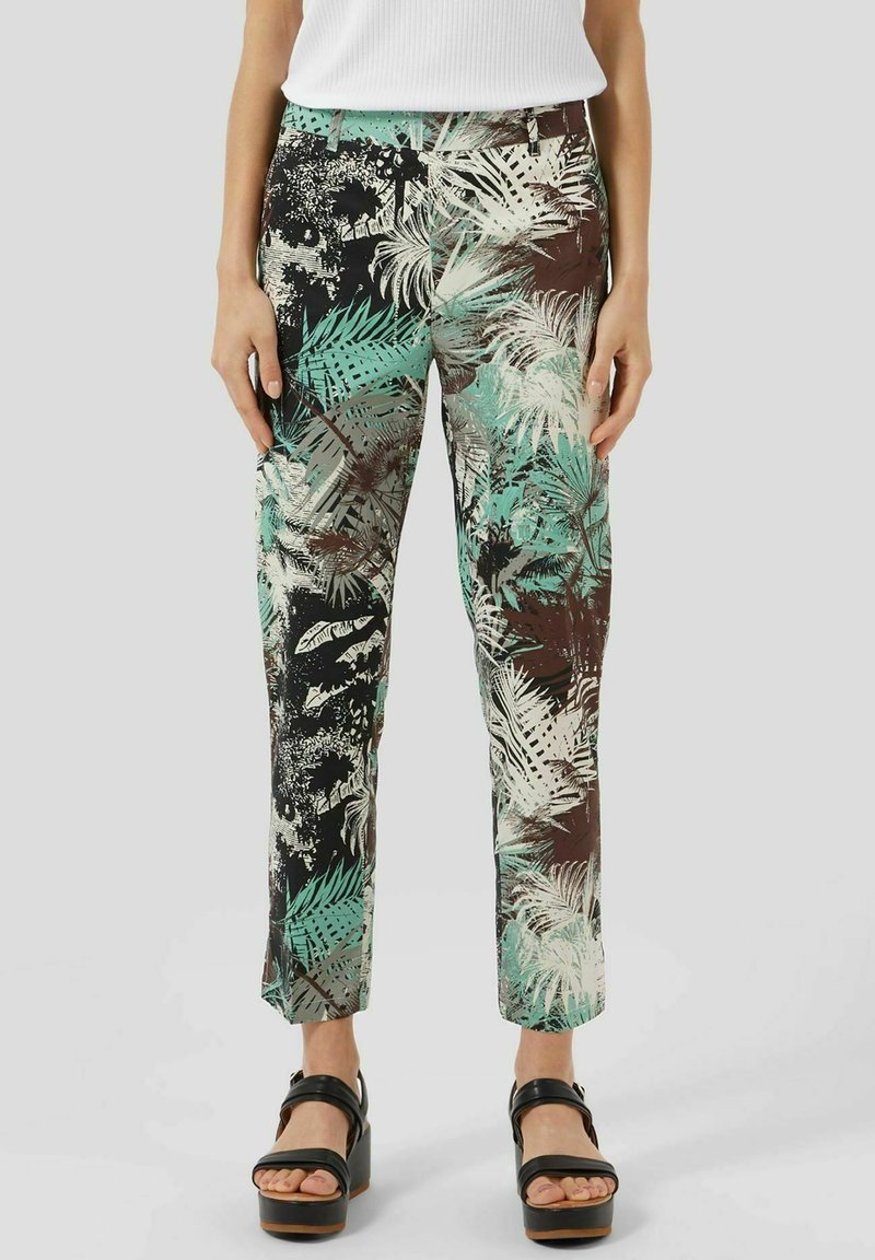 Femme portant un pantalon slim imprimé tropical dans des tons verts, marron et crème, avec des sandales plateformes noires et un top blanc sans manches.