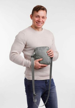 Šedá křížová taška s texturovaným povrchem, plochým designem, nastavitelným popruhem a zipovým uzávěrem, kterou drží osoba v neformálním oblečení.