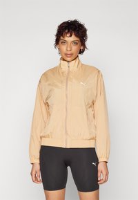Veste légère de couleur pêche avec fermeture éclair, col haut, poignets élastiques et ourlet élastiqué. Présente un logo Puma blanc sur la poitrine.