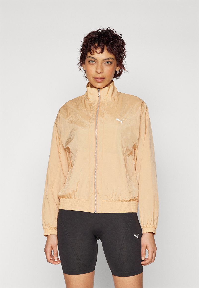 Veste légère de couleur pêche avec fermeture éclair, col haut, poignets élastiques et ourlet élastiqué. Présente un logo Puma blanc sur la poitrine.