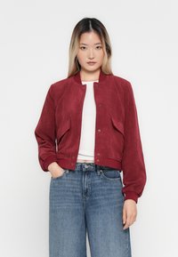 Young woman with long hair wears a red corduroy bomber jacket over a white top and blue jeans, hands partially in pockets, against white background.