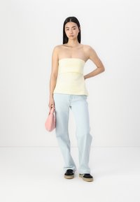 Żółta bezramkowa bluzka z fasonem peplum, połączona z jasnoniebieskimi dżinsami i czarnymi sneakersami. Trzyma różową torebkę z materiału. Gładka tekstura.