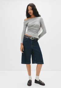 Graues Langarm-Oberteil mit schulterfreiem Schnitt, blaue Jeans-Bermuda-Shorts, gemusterter Gürtel und dunkle Mokassins, getragen mit hellen Socken. Neutraler Hintergrund.