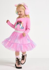 Abito lungo a maniche lunghe di colore rosa con un design di paillettes arcobaleno, gonna in tulle a strati e una cintura glitterata. Abbinato a stivaletti rosa con lacci.