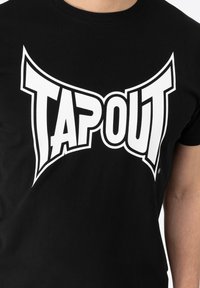 Musta puuvillapaita, jossa on suuri valkoinen "TAPOUT" -logo. Paidassa on klassinen pyöreä kaula-aukko ja lyhyet hihat. Sujuva kangas.